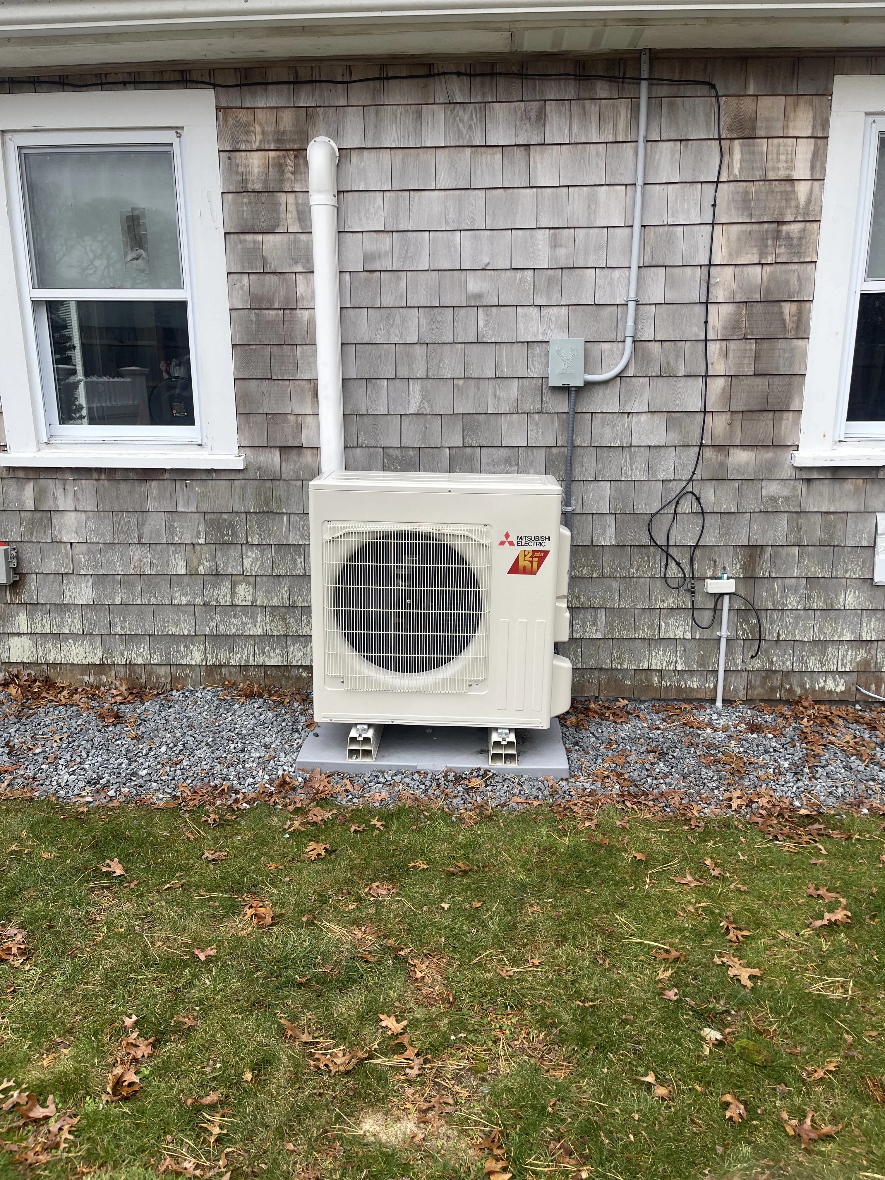 Mitsubishi heat pump outdoor unit installed on concrete pad against cedar shake siding with white trim windows